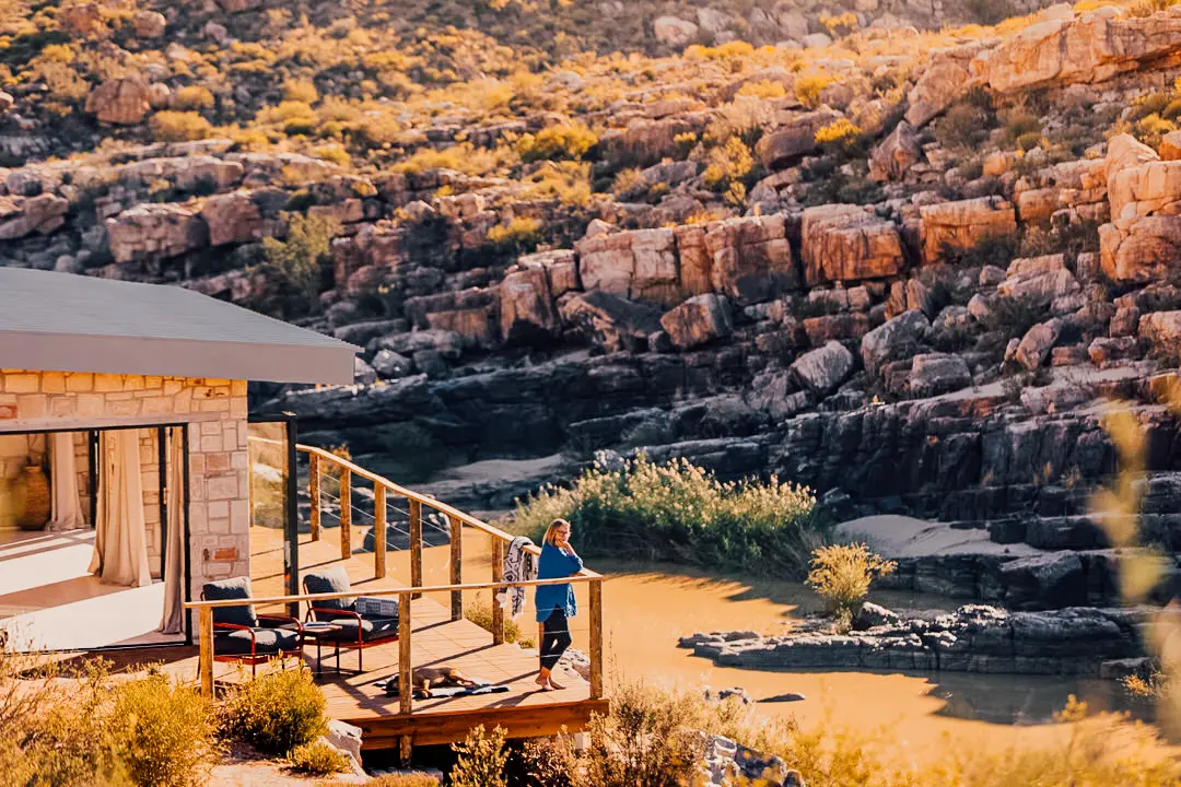 Woman standing on wooden deck of stone chalet at Bliss and Stars, Doring River and layered Cederberg rock face behind, golden afternoon light