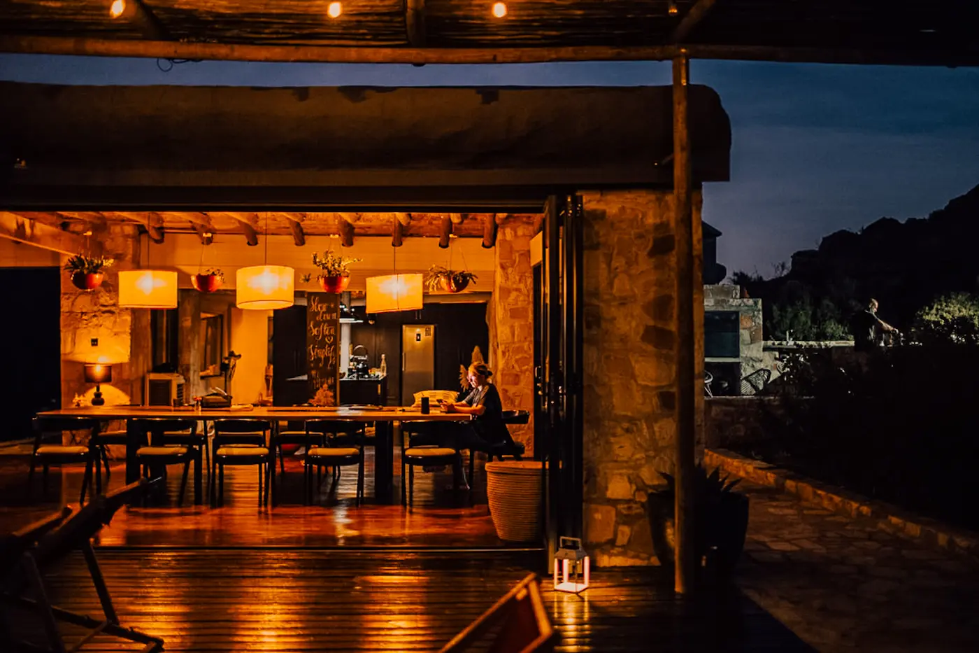Long communal dining table under woven pendant lights at night, one guest seated at the far end, an open deck and the dark Cederberg beyond.