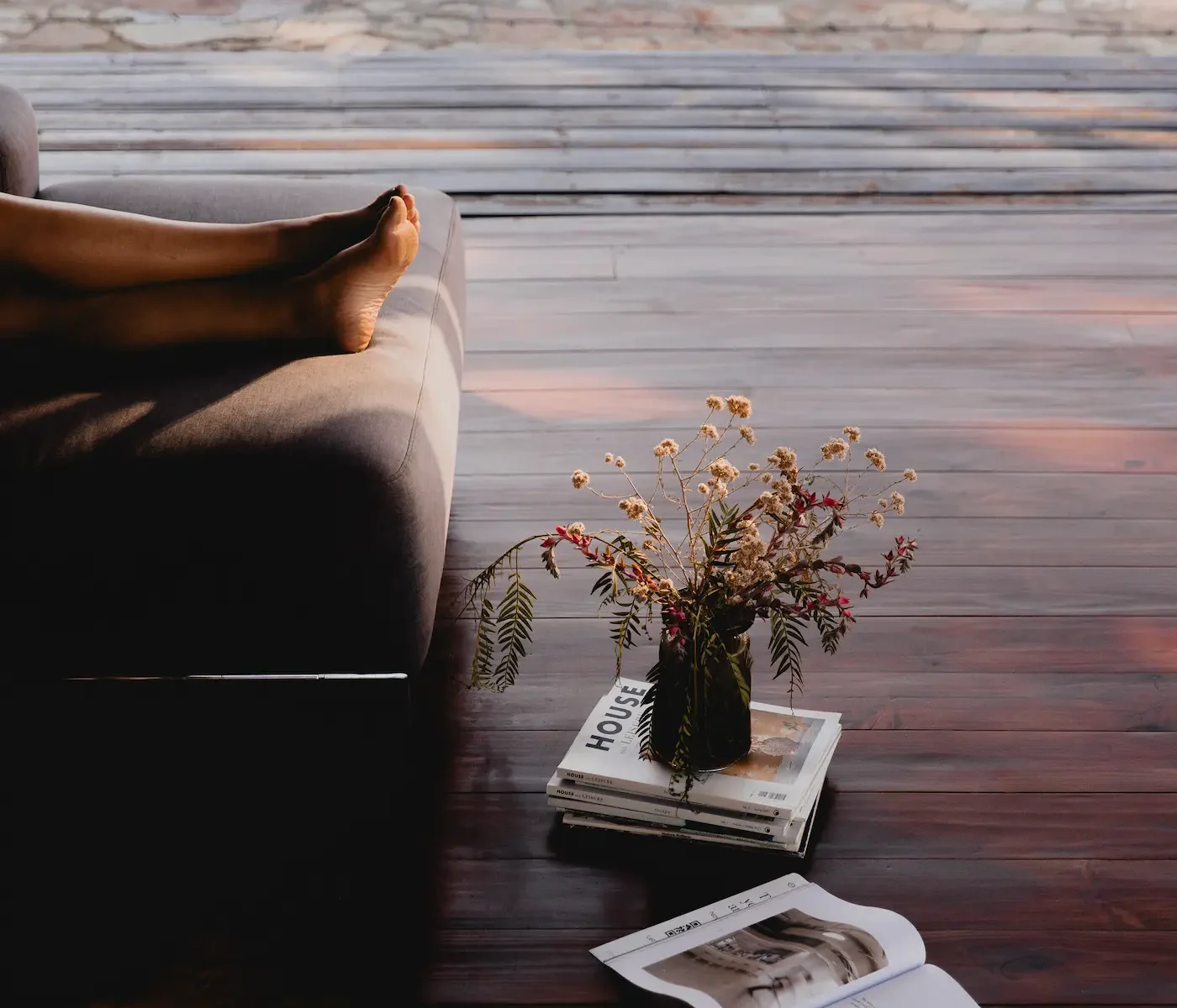 Bare feet resting on sofa arm on wooden terrace, open magazine and dried wildflowers on floor, warm evening light, Cederberg rock view through glass