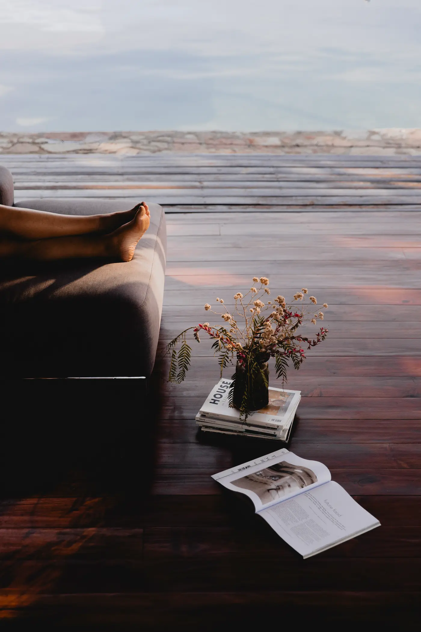 Bare feet resting on sofa arm on wooden terrace, open magazine and dried wildflowers on floor, warm evening light, Cederberg rock view through glass