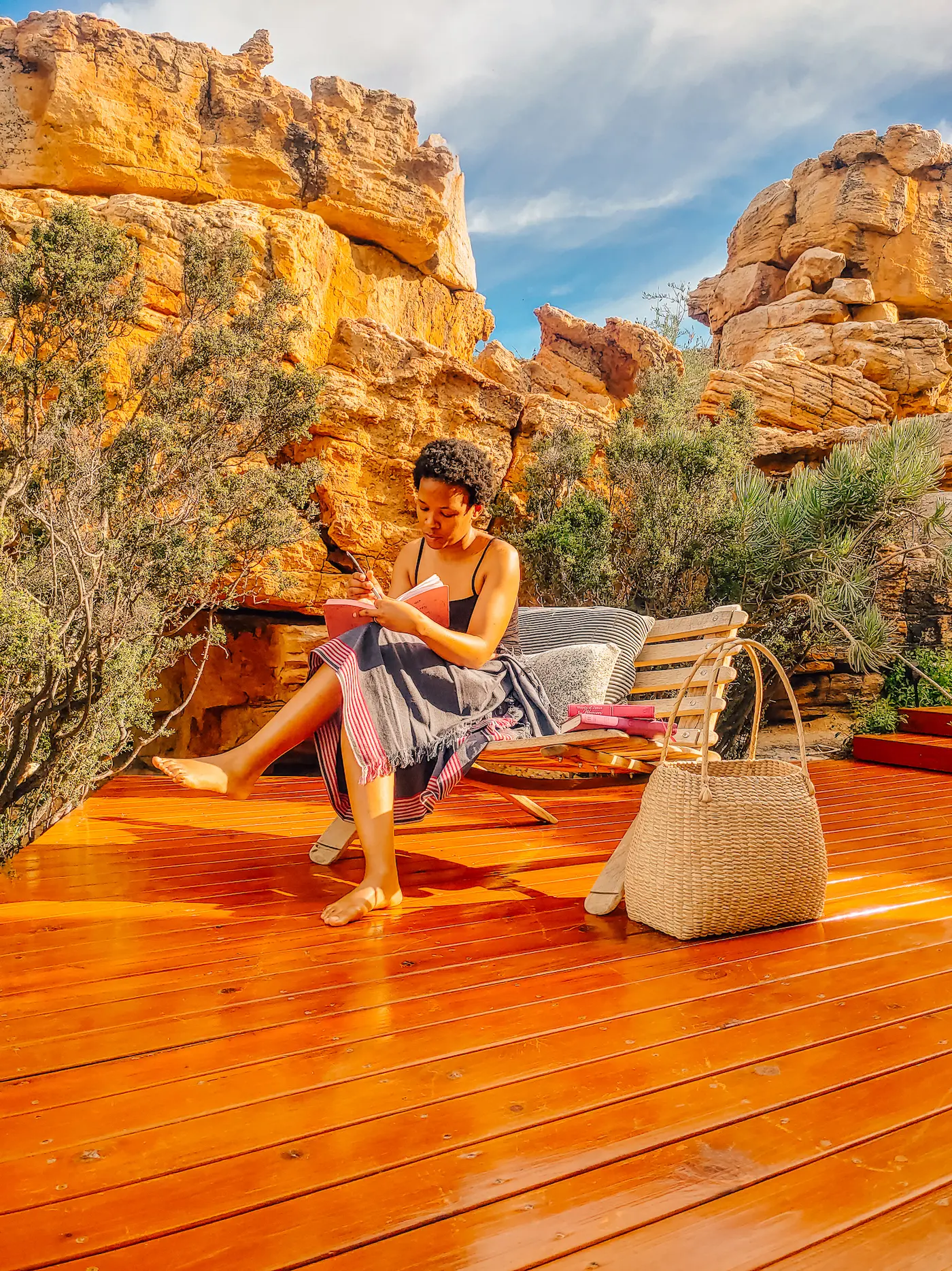 Woman sitting barefoot on wooden deck writing in journal, ochre Cederberg sandstone rocks behind, Unmade women's retreat