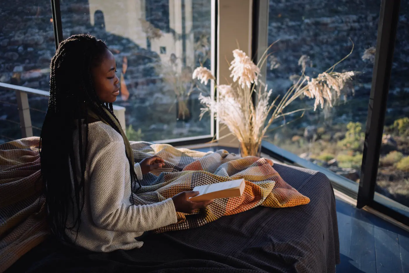 A woman wrapped in a blanket reads a book beside floor-to-ceiling windows framing the rocky Cederberg landscape